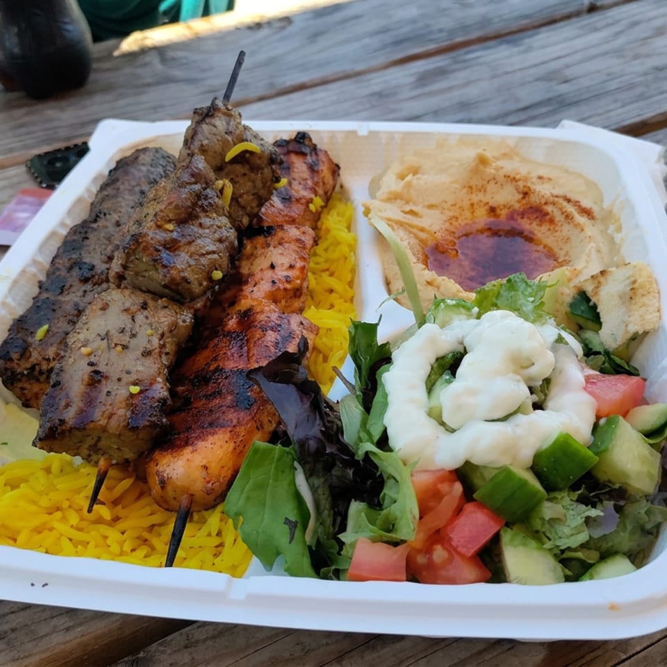 Platter of grilled meat skewers, yellow rice, hummus, and salad served in a takeaway tray on a wooden table.