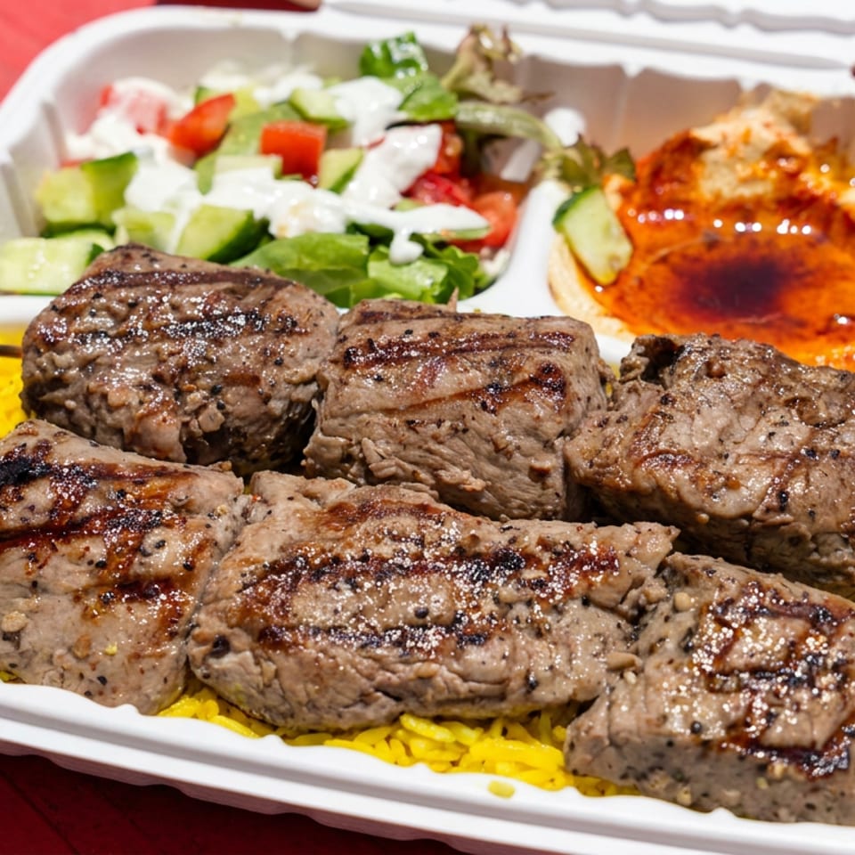 Grilled meat skewers served over yellow rice with salad and hummus in a white takeout container.
