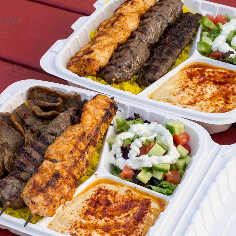 Two takeout trays of grilled kebabs and meat, yellow rice, salad, and hummus on a red picnic table.